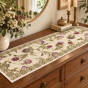 Puede incluir: Un camino de mesa rectangular de tela color crema con un patrón botánico repetitivo de alcachofas y cardos morados y verdes, enmarcado por un borde tejido de color verde oscuro con intrincados diseños de volutas.