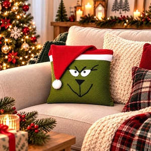Könnte beinhalten: Ein quadratisches, grünes Strickkissen mit einem aufgestickten Gesicht, bestehend aus weißen Augen sowie einer schwarzen Nase und einem Mund. Das Kissen ist mit einer roten Strick-Weihnachtsmütze mit weißem Rand und weißem Bommel versehen.