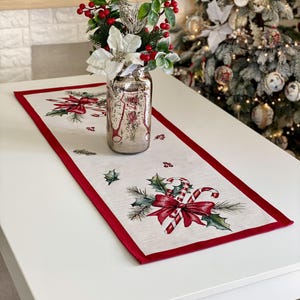 May include: A rectangular table runner featuring a cream-colored fabric with a red border. The runner is decorated with printed illustrations of candy canes, red bows, green holly leaves, and pine needles. A glass vase sits on the runner.