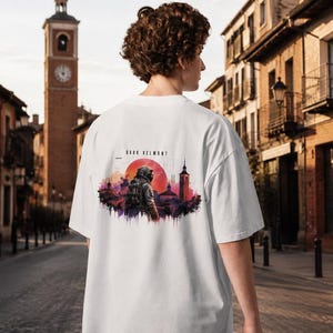 Puede incluir: Una camiseta blanca con un estampado gráfico en la espalda. El diseño muestra una figura con casco frente a un gran sol rojo, una torre alta y un paisaje urbano. El texto "DARK VELMONT" está impreso en negro sobre la ilustración central.