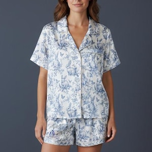 Op de afbeelding: Een tweedelige pyjamaset bestaande uit een shirt met korte mouwen en een short. De set is wit met een blauwe bloemen- en konijnenprint. Het shirt heeft een klassieke kraag en is gemaakt van een zachte, lichte stof.