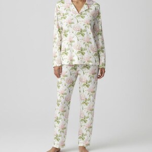 May include: A two-piece pajama set featuring a floral pattern. The set includes a long-sleeved top with a collared neckline and pants. The pajamas are white with a repeating pattern of pink roses and green leaves.