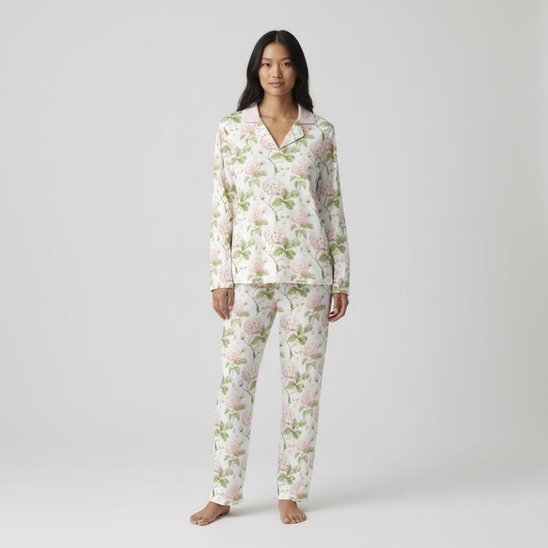 May include: A two-piece pajama set featuring a floral pattern. The set includes a long-sleeved top with a collared neckline and pants. The pajamas are white with a repeating pattern of pink roses and green leaves.