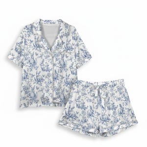 Op de afbeelding: Een tweedelige pyjamaset in wit en blauw met een toile de Jouy-patroon met konijnen en bloemen. De set bestaat uit een overhemd met korte mouwen en bijpassende shorts. De shorts hebben een taille met trekkoord.