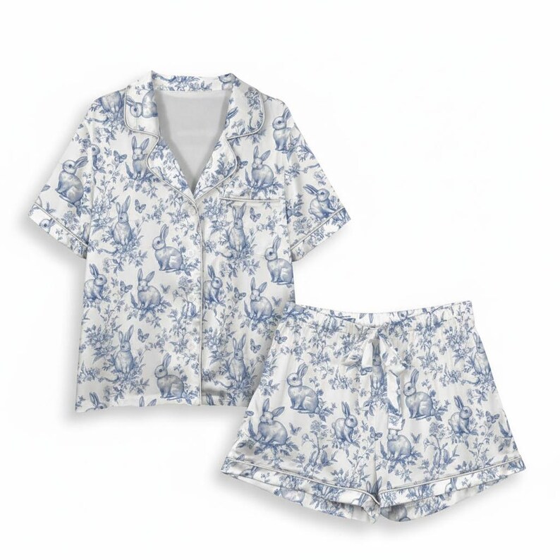 Op de afbeelding: Een tweedelige pyjamaset in wit en blauw met een toile de Jouy-patroon met konijnen en bloemen. De set bestaat uit een overhemd met korte mouwen en bijpassende shorts. De shorts hebben een taille met trekkoord.