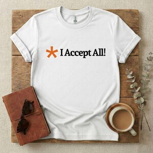 May include: A white short-sleeved t-shirt featuring an orange asterisk symbol followed by the black text "I Accept All!" printed across the centre of the chest. The garment has a crew neckline and a standard fit with visible stitching details.