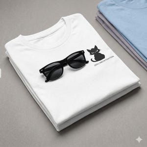 Puede incluir: Camiseta blanca con un gato negro y el texto "ZERO MEOWTIVATION". Gafas de sol negras sobre la camiseta doblada. Se ven camisetas dobladas adicionales en azul claro y gris.