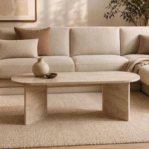 Puede incluir: Una mesa de centro ovalada con un acabado en tono beige claro similar a la piedra, con una superficie lisa sostenida por dos patas verticales anchas y planas. La mesa presenta una apariencia texturizada y moteada sobre una alfombra tejida.
