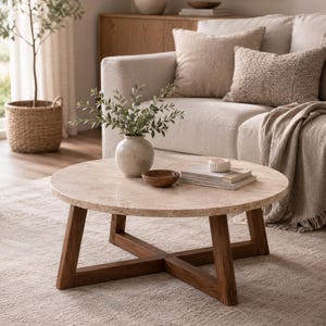 Puede incluir: Una mesa de centro redonda con una tapa de piedra clara y patas de madera oscura en forma de X. La mesa está en un salón con un sofá beige, cojines y una alfombra. Un jarrón con vegetación y un cuenco de madera pequeño están sobre la mesa.