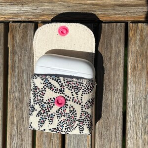 Puede incluir: Una funda de tela color crema con un estampado floral de puntos negros, rosas y azules, que contiene una funda blanca para auriculares. La funda tiene un cierre de botón a presión rosa y un botón a presión rosa a juego en la solapa. La funda está abierta, revelando los auriculares.