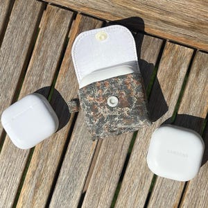 Puede incluir: Una funda estampada con interior blanco y cierre de botón a presión, junto a dos fundas blancas para auriculares. Una funda está cerrada, la otra está abierta. El logotipo de Samsung es visible en una funda. Los artículos están sobre una superficie de madera.