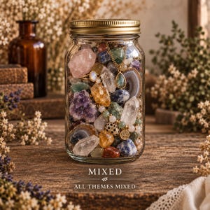 Op de afbeelding: Een glazen pot met een gouden deksel gevuld met een verscheidenheid aan kleurrijke ruwe edelstenen, kristallen en sieraden, waaronder een gouden ketting met een druppelvormige hanger en een boombedel. De tekst MIXED ALL THEMES MIXED is zichtbaar.