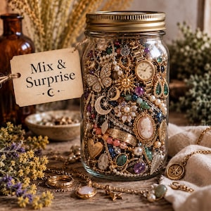 May include: A clear glass jar with a gold lid filled with assorted jewelry including necklaces, bracelets, a watch, a cameo, and charms. A beige paper tag attached to the jar features the text Mix & Surprise with a crescent moon and star symbol.