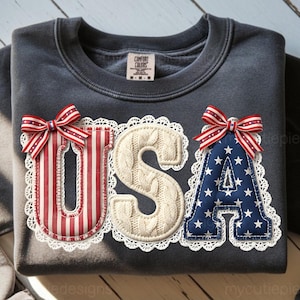 May include: Dark gray sweatshirt with the word "USA" appliqued on the front. The letters are decorated with red and white stripes, a cream-colored knit pattern, and blue stars. Each letter is trimmed with white lace and topped with a red, white, and blue bow.