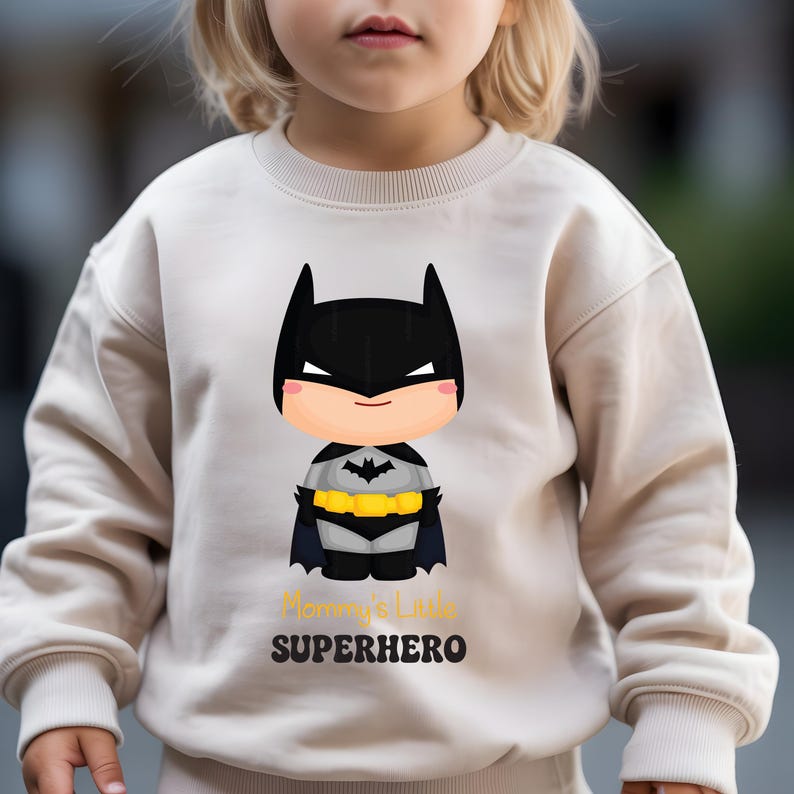 May include: Beige sweatshirt with a cartoon Batman design. The character has a black cowl, cape, and yellow utility belt. The text "Mommy's Little SUPERHERO" is printed below. The sweatshirt is designed for children.