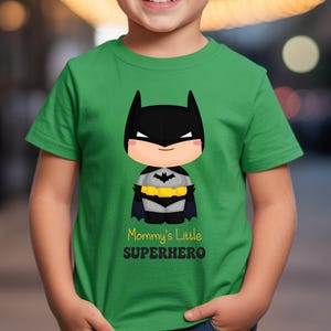 May include: A green t-shirt with a cartoon Batman design. The design features a black mask, cape, and utility belt. The text "Mommy's Little Superhero" is printed below the character.