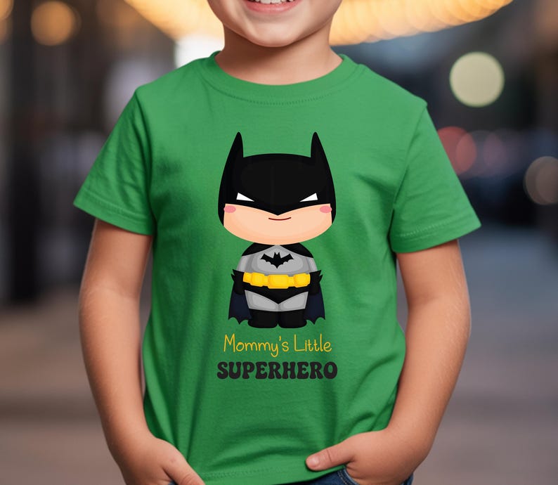 May include: A green t-shirt with a cartoon Batman design. The design features a black mask, cape, and utility belt. The text "Mommy's Little Superhero" is printed below the character.
