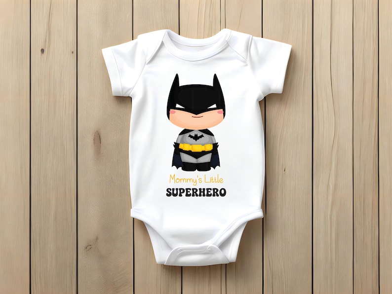 May include: White baby bodysuit with a cartoon Batman design. The illustration shows Batman with a black cowl, cape, and yellow utility belt. The text "Mommy's Little SUPERHERO" is printed in yellow and black. The background is wooden.