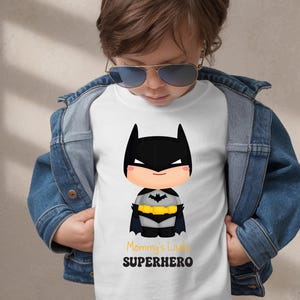 May include: White t-shirt with a cartoon Batman design, including a black cowl, cape, and yellow belt. The text "Mommy's Little SUPERHERO" is printed below. The shirt is worn with a denim jacket.