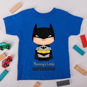 May include: Royal blue t-shirt with a Batman cartoon graphic, including a black mask and cape, and a yellow belt. The text reads "Mommy's Little Superhero". Toy cars and wooden blocks are scattered around the shirt.