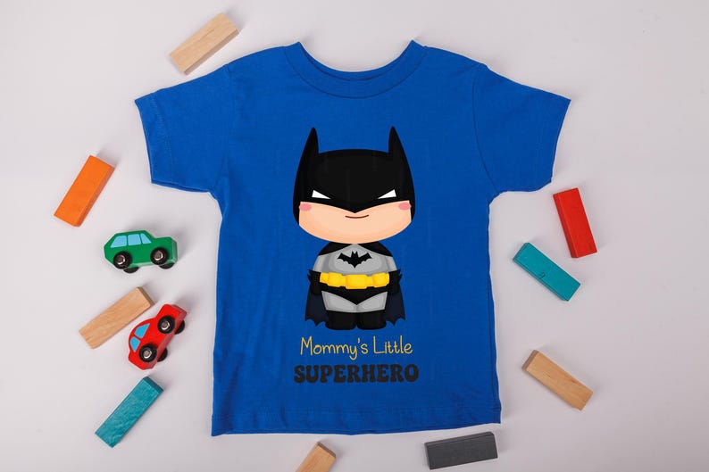 May include: Royal blue t-shirt with a Batman cartoon graphic, including a black mask and cape, and a yellow belt. The text reads "Mommy's Little Superhero". Toy cars and wooden blocks are scattered around the shirt.