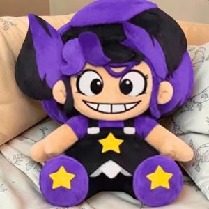 May include: A plush doll with a cartoon-style design. The doll has a purple and black hairstyle, large eyes, and a wide smile. It wears a black outfit with yellow star accents on the chest and legs. The doll is sitting on a patterned pillow.