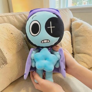 May include: A soft toy doll with a light blue face, a black eye patch, and a white cross. The doll has a purple hat and cape, and a blue star-shaped body. The doll is being held by a person.