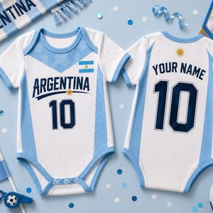 Puede incluir: Dos bodies para bebé blancos y azul claro con el texto ARGENTINA y el número 10 en la parte delantera, y el texto YOUR NAME y el número 10 en la parte trasera. Los diseños incluyen un pequeño emblema de la bandera argentina y un sol.