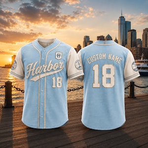 Op de afbeelding: Een lichtblauw en crèmekleurig honkbalshirt met het woord Harbor in script, het nummer 18 en een embleem op de voorzijde. De achterzijde toont de tekst Custom Name boven het nummer 18. Beide mouwen zijn voorzien van een rond vuurtorenlogo.