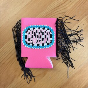 May include: A pink drink koozie with black fringe on the sides. The koozie has an embroidered patch with the word "PARTY" in a checkered pattern, outlined in blue and white.