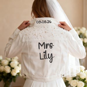 Puede incluir: Una chaqueta vaquera blanca con el cuello bordado con la fecha 09.08.26. La parte posterior de la chaqueta está decorada con perlas blancas dispersas y el texto bordado en negro Mrs Lily. La chaqueta se lleva con un velo blanco transparente.