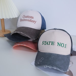 Puede incluir: Gorras de béisbol con viseras desgastadas. Una gorra tiene "Custom Embroidery" bordado en marrón, otra dice "STATE NO.1" en verde. Las gorras tienen un diseño de dos tonos con una corona blanca y una visera azul o gris.