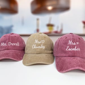 Puede incluir: Se muestran tres gorras de béisbol. Dos son de color rosa polvoriento y una es de color beige. Cada gorra tiene texto bordado en blanco con los nombres "Mrs. Duvall", "Mrs. Chumley" y "Mrs. Escobar".