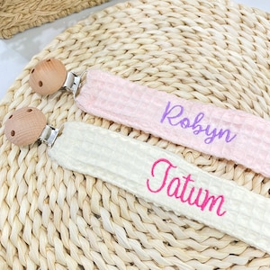 Puede incluir: Dos clips para chupete de bebé con cuentas de madera y cierres metálicos. Uno es rosa claro con el nombre "Robyn" bordado en morado. El otro es blanquecino con "Tatum" en rosa. Ambos están sobre una superficie tejida.