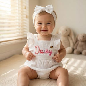 Könnte beinhalten: Ein lächelndes Baby trägt einen weißen Strampler mit dem Namen "Daisy" in rosa Schrift gestickt. Der Strampler hat Rüschenärmel und Träger. Ein passendes weißes Schleifen-Stirnband vervollständigt das Outfit. Eine kleine gestickte Biene befindet sich neben dem Namen.