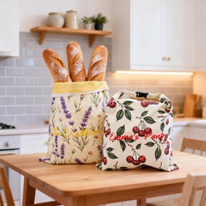 Puede incluir: Dos bolsas de tela sobre una mesa de madera. La bolsa más alta tiene un estampado de lavanda y margaritas con el texto "The William" y contiene tres baguettes. La bolsa más corta tiene un estampado de cerezas con el texto "Emma's Pantry".