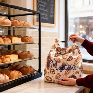 May include: A cream-colored fabric lunch bag featuring a repeating pattern of various breads and geometric motifs, with the name Mateo embroidered in blue script across the front and a black plastic buckle closure at the top handle.
