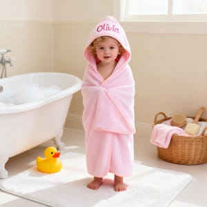 May include: A soft, light pink hooded towel featuring the name Olivia embroidered in a darker pink script on the hood. The towel is wrapped around a small child, providing full coverage with a simple, clean design and finished edges.