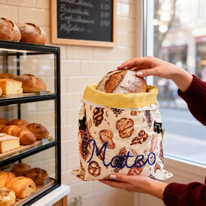 Puede incluir: Una bolsa de pan de tela color crema con un patrón repetitivo de varios productos horneados y el nombre Mateo bordado en letra azul. La bolsa tiene un forro interior amarillo y hebillas de plástico negro en los laterales.
