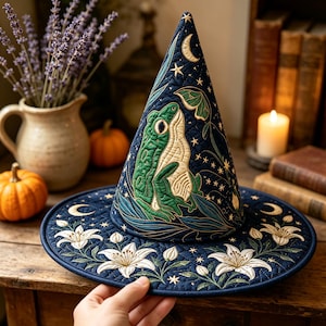 Puede incluir: Un sombrero cónico acolchado de color azul oscuro con una rana verde bordada, flores de lirio blancas, una luna creciente y pequeñas estrellas. La tela tiene una textura pespunteada, con un ala ancha y una punta superior con bordados detallados.