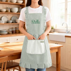 May include: A sage green and white striped apron featuring ruffled shoulder straps, a pleated hem, and a large front pocket. The chest area is embroidered with the name Stella in a dark green cursive script, and a small rectangular label is attached.