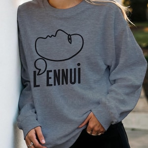 May include: A gray sweatshirt with the word "L'ENNUl" in black, along with a black line drawing of a face. The sweatshirt is a casual style, suitable for everyday wear.