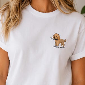 Può includere: T-shirt bianca con un cane dei cartoni animati che tiene un coltello. Il cane è marrone chiaro con occhi neri e naso nero. Il testo "I REQUIRE SNACKS" è stampato sotto.