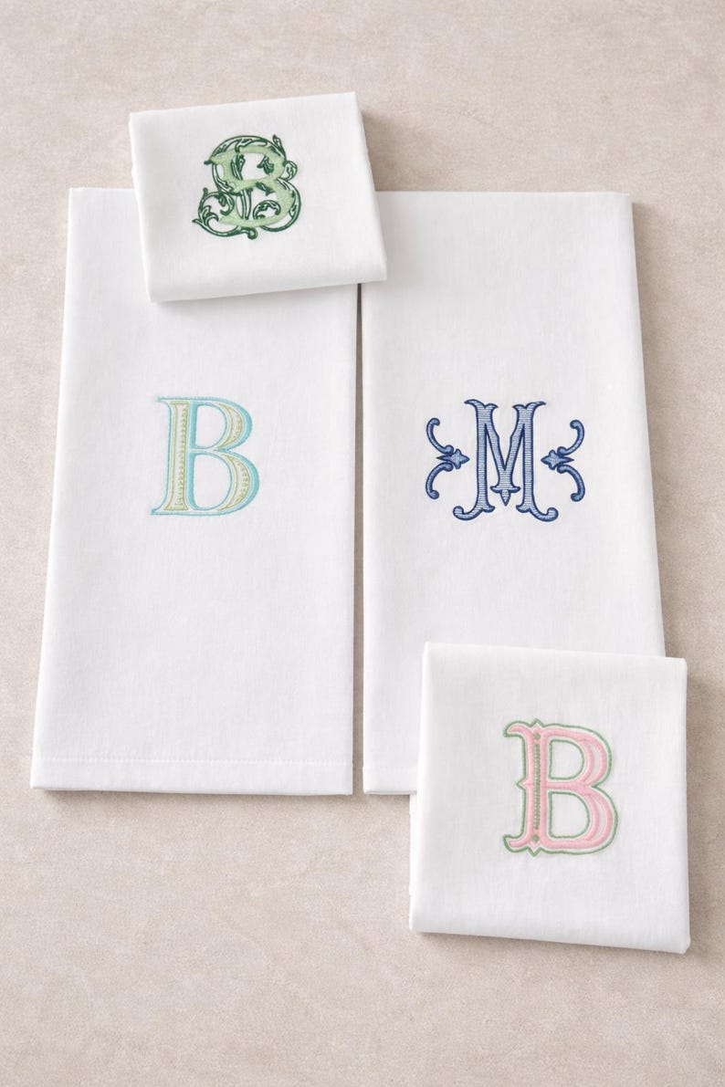 May include: White embroidered towels and napkins with various initial letters. The letters are in shades of blue, green, and pink. The towels are rectangular, and the napkins are square.