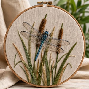May include: A circular embroidery piece in a wooden hoop. It depicts a dragonfly with blue and teal detailing, set amongst embroidered cattails and reeds. The dragonfly's wings are transparent, and the reeds are in shades of green and tan. The background is a beige fabric.