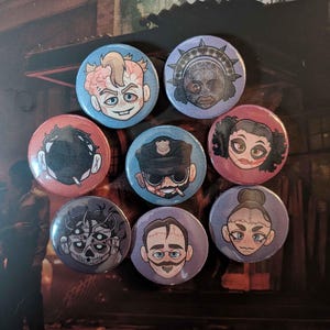 Op de afbeelding: Een verzameling van zeven ronde buttons met speldsluiting en gestileerde karakterillustraties. De ontwerpen bevatten een figuur met blootliggende hersenen, een personage met een stekelige kroon, een rokende figuur, een politieagent, een clownachtig gezicht, een schedel met meerdere ogen en een man met een snor.