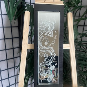 Może przedstawiać: Prostokątne lustro z czarną ramą, z wytrawionym białym wzorem smoka. Smok wije się po powierzchni lustra. Lustro jest prezentowane na drewnianym sztaludze, z zielenią w tle.