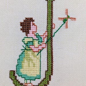 Op de afbeelding: Een kruissteekborduurwerk op witte stof met de letter J in het groen met een bruine omlijning. Een figuur met bruin haar, een lichtgroene jurk en een geel schort staat naast de letter en houdt een groen touwtje vast dat verbonden is met een kleine roze windmolen.