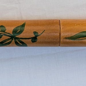 Op de afbeelding: Een cilindervormige houten koker met een gladde, lichtbruine afwerking, voorzien van handgeschilderde rode bloemen met donkere harten en groene bladeren die over het oppervlak verspreid zijn. De koker is verdeeld in twee delen door een naad.