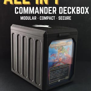 May include: A black modular deck box with a clear front display window containing a Captain America trading card. The box features vertical ribbed side panels and a top lid with a white logo. Text reads ALL IN 1 COMMANDER DECKBOX MODULAR COMPACT SECURE FITS 100+ DOUBLE SLEEVED CARD DICES & TOKENS.
